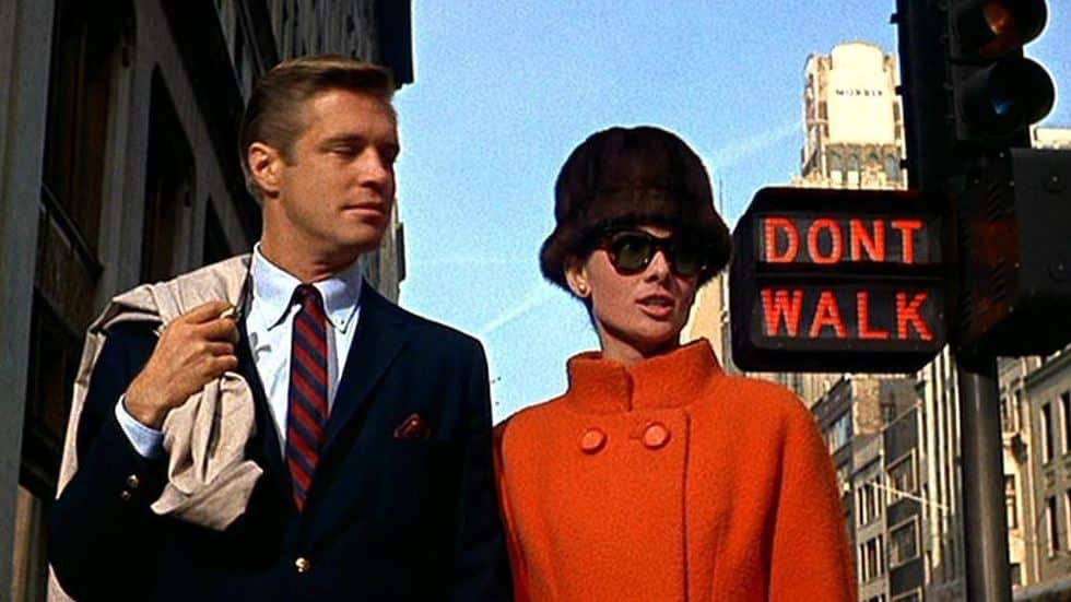 Audrey Hepburn con cappotto arancione, cappello in pelliccia e occhiali da sole scuri accanto a George Peppard